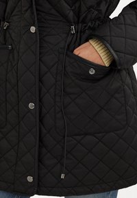 Svart vadderad jacka med åtdragen midja, med två framfickor, metallknappar och en slät, matt yta.