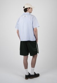 Jeune homme portant une chemise à manches courtes rayée bleu clair, un short rayé noir, des chaussettes blanches, des chaussures noires et une casquette de baseball blanche, vu de dos.