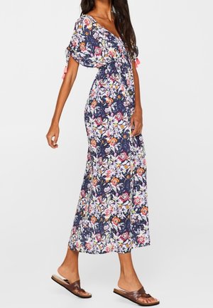 Vrouw in een marineblauwe maxi-jurk met een kleurrijk bloemenpatroon, vliegermouwen en bruine instapsandalen, staand tegen een effen achtergrond.