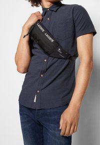 Osoba oblečená v námořnickém modrém krátkém rukávu košili a džínech, držící černou crossbody tašku Lacoste přes hruď.