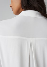 Witte blouse met een klassieke kraag, een gladde textuur en een plooidetail aan de achterkant. De stof is lichtgewicht en iets doorschijnend.