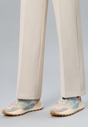 Pantalon beige porté avec des baskets pastel rose, bleu et crème, dotées de semelles texturées et d'un design à lacets, sur un fond gris.