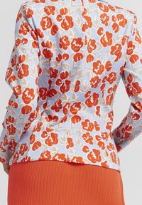 Veste à motif floral avec des fleurs rouges et beiges sur un fond bleu clair. Design ajusté avec une texture de tissu lisse.