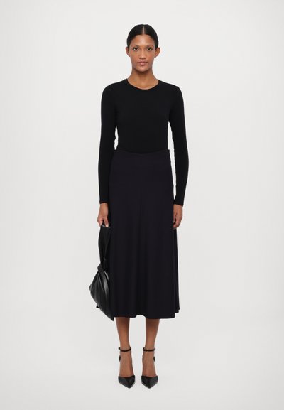 Top negro de manga larga con cuello redondo, combinado con una falda midi oscura y vaporosa. La modelo lleva un bolso negro y unos tacones negros.