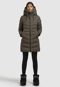 Olive green puffer coat with a high collar, zip closure, and two side pockets. Worn with black leggings and black boots. Smooth texture, quilted design.