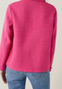 Blazer strutturato rosa con un design aderente, maniche lunghe e colletto. Presenta un dettaglio a bottone sui polsini. Indossato sopra jeans in denim blu.