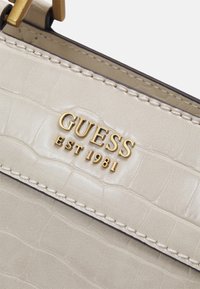 Beige, tekstureret kunstlæderhåndtaske med krokodillemønster. Har gyldent "GUESS EST 1981"-logo og kontrastfarvet syning nær kanten.