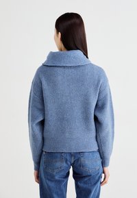 Blå strikket genser med sjalkrage, med ribbestruktur og løs passform. Kombinert med klassiske blå jeans, sett fra baksiden.