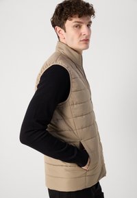 Μπεζ γιλέκο με ψηλό γιακά, οριζόντιο καπιτονέ σχέδιο και πλαϊνές τσέπες, φορεμένο πάνω από μαύρο πουκάμισο με μακριά μανίκια.