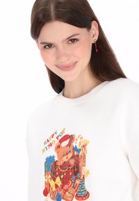 Fehér pulóver, amelyen egy színes rajzfilmmedve ajándékokkal, piros kalapban látható, valamint a "HAPPY MY MO POP" felirat. Puha anyagból, kerek nyakkivágással.