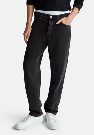 Uomo che indossa jeans neri sbiaditi a vestibilità ampia con cinque tasche, fascia bianca visibile, maglia nera a maniche lunghe e scarpe da ginnastica bianche, in piedi con una mano in tasca.