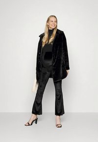 Casaco de pêlo preto sintético com gola, usado por cima de um top preto e calças flare combinando. A modelo leva uma mala branca texturada.