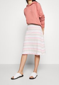 Rosa hoodie med vit logotyp, ihopparad med en knä-lång randig kjol i pastellfärger. Skorna inkluderar vita slidesandaler med svarta sulor.