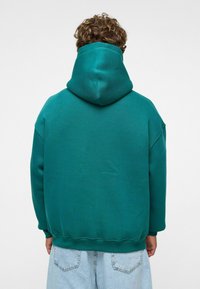 Turquoise hoodie met een grote capuchon, ontspannen pasvorm, ribgebreide manchetten en zoom; zachte textuur, gedragen met lichtblauwe baggy jeans.