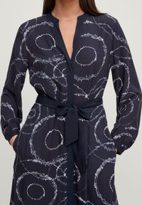 Blusa navy con motivi circolari bianchi, maniche lunghe, scollatura a V e dettaglio in vita annodato. Realizzata in tessuto leggero.