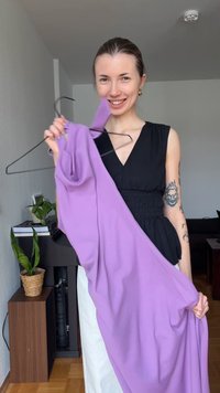 Γυναίκα που χαμογελάει μέσα σε εσωτερικό χώρο, κρατώντας ένα μωβ φόρεμα σε μαύρη κρεμάστρα, φοράει μαύρο αμάνικο τοπ και λευκό παντελόνι, με τατουάζ στα χέρια.