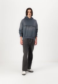 Blauer Kapuzenpullover mit Kordelzug und Fronttasche, mit dem Logo "PROJECT CORE" in dunkler Schrift. Getragen mit schwarzen Jeans und weißen Sneakers.