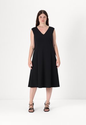 Svart A-linje-kjole med V-hals, uten ermer, laget av mykt stoff. Kombinert med svarte sandaletter med stropp. Enkel og elegant stil.