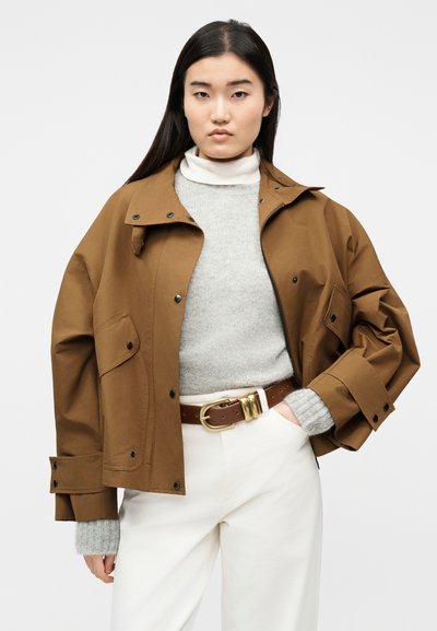 Γυναίκα με μακριά σκούρα μαλλιά, φοράει ένα καφέ φαρδύ μπουφάν πάνω από γκρι πουλόβερ και άσπρο παντελόνι με καφέ ζώνη, στέκεται μπροστά σε απλό φόντο.