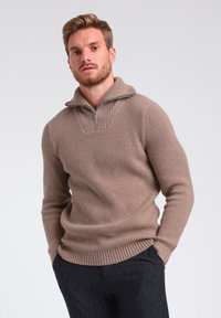 Beige gestrickter Pullover mit einem halben Reißverschlusskragen, der vertikale Rippenstrukturen und eine lockere Passform aufweist. Getragen mit dunkelgrauen, strukturierten Hosen.