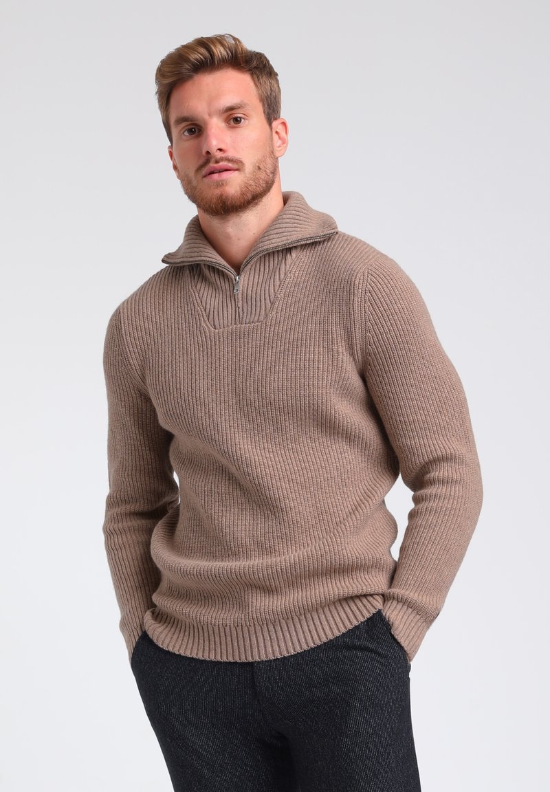 Beige gestrickter Pullover mit einem halben Reißverschlusskragen, der vertikale Rippenstrukturen und eine lockere Passform aufweist. Getragen mit dunkelgrauen, strukturierten Hosen.