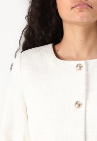 Vit texturerad jacka med rund halsringning och guldbutiksdetaljer framtill. Tyget verkar vara vävt och mjukt.