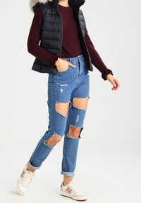 Svart pufferväst med pälsluva, vinröd långärmad tröja, slitna blå jeans med flera stora urklipp, och vita sneakers.