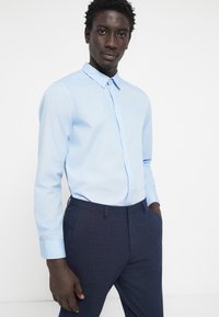 Svijetloplava pamučna košulja s ovratnikom na gumbe, dugim rukavima i glatkom teksturom, uparen s tamnoplavim hlačama s uzorkom.