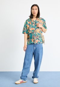 Short-sleeve shirt in green with floral patterns, paired with loose blue denim jeans and white sneakers with red stripes.
