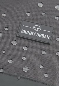 Țesătură neagră texturată cu picături de apă circulare ridicate și o etichetă rectangulară neagră cu inscripția "JOHNNY URBAN" în litere albe.