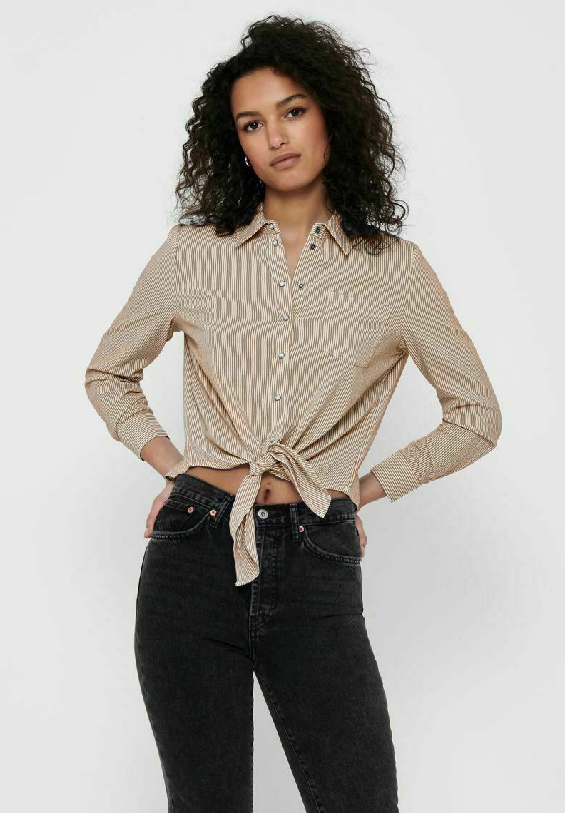 Striped button-up shirt in beige and white, featuring long sleeves, a front tie, and a chest pocket, paired with black high-waist jeans.