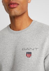 Grijze sweatshirt met een geribbelde halslijn, met een geborduurd "GANT"-logo en een schildontwerp in rood, wit en blauw op de borst.