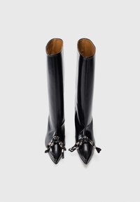 Botas negras de cuero liso, de caña alta y punta afilada, con herrajes decorativos y borlas en la puntera como detalle.