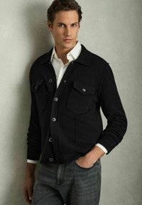 Zwarte wolblend cardigan met vier frontzakken, knoopsluiting en geribde manchetten, in lagen over een wit shirt en grijze jeans.