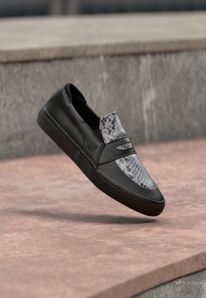 Sort lædersko med slip-on-design og gråt slangeskindsmønster på overdelen, svævende over en trinopdelt betonoverflade.