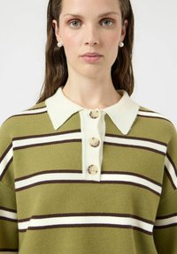 Kvinne iført grønn, hvit og brunstripet genser med hvit krage og tre store knapper, vendt fremover mot en ensfarget bakgrunn.