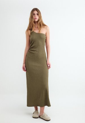 Junge Frau mit langen Haaren, die ein olivgrünes, strukturiertes One-Shoulder-Kleid und beige Slipper trägt, steht vor einem schlichten weißen Hintergrund.