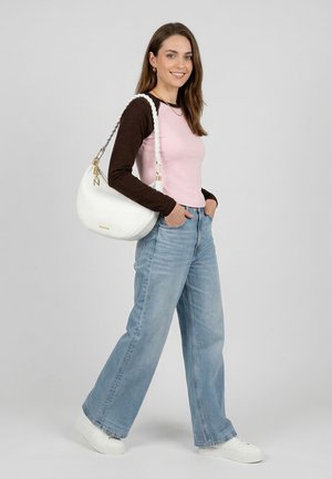 Joven sonriente, vestida con una camiseta de manga larga rosa y marrón, jeans azul claro de pierna ancha, zapatillas blancas y llevando un bolso blanco de hombro.