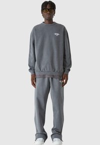 Szürke kapucnis pulóver és hozzá illő, puha anyagú melegítőnadrág. Az elején egy kis logó található, és laza szabás jellemzi. Fehér sportcipőkkel egészíti ki a megjelenést.