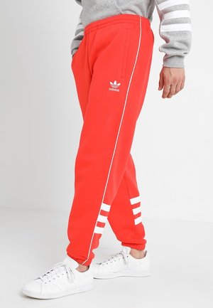 Personne portant un pantalon de survêtement Adidas rouge vif avec des bandes blanches près des chevilles et un logo, associé à des baskets blanches et un sweat-shirt gris.