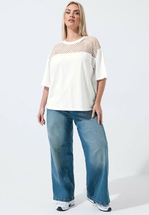 Mujer rubia con una blusa blanca con paneles de malla, jeans azules de pierna ancha y zapatillas blancas, de pie frente a un fondo claro y liso.