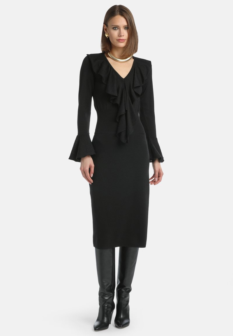 Czarna dzianinowa sukienka z falbaniastym dekoltem, dopasowanym górnym partem i rozszerzającymi się rękawami, w połączeniu z czarnymi kozaczkami na obcasie do kolan.