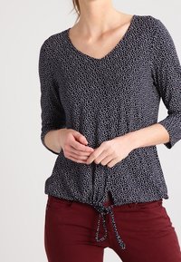 Námořnická blůza s bílým abstraktním vzorem, výstřih do V, rukávy 3/4 a stahovací lem. Přiřazena k vínovým úzkým kalhotám.