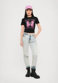 Kvinne som har på seg en svart crop top med rosa sommerfugl og tekst, lysvaskede jeans, svarte støvler og en rosa baseballcaps, stående mot en hvit bakgrunn.