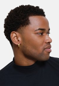 Gold hoop earring with a textured, multi-layered design. Worn on a person with short, curly hair, in a black turtleneck shirt.