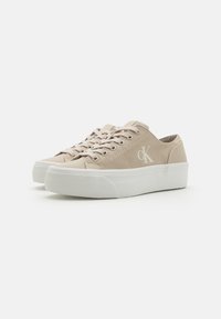 Beige Leinensneaker mit weißen Gummisohlen, Schnürdesign und auffälligem "CK"-Logo auf der Seite. Glatte Textur, lässiger Stil.