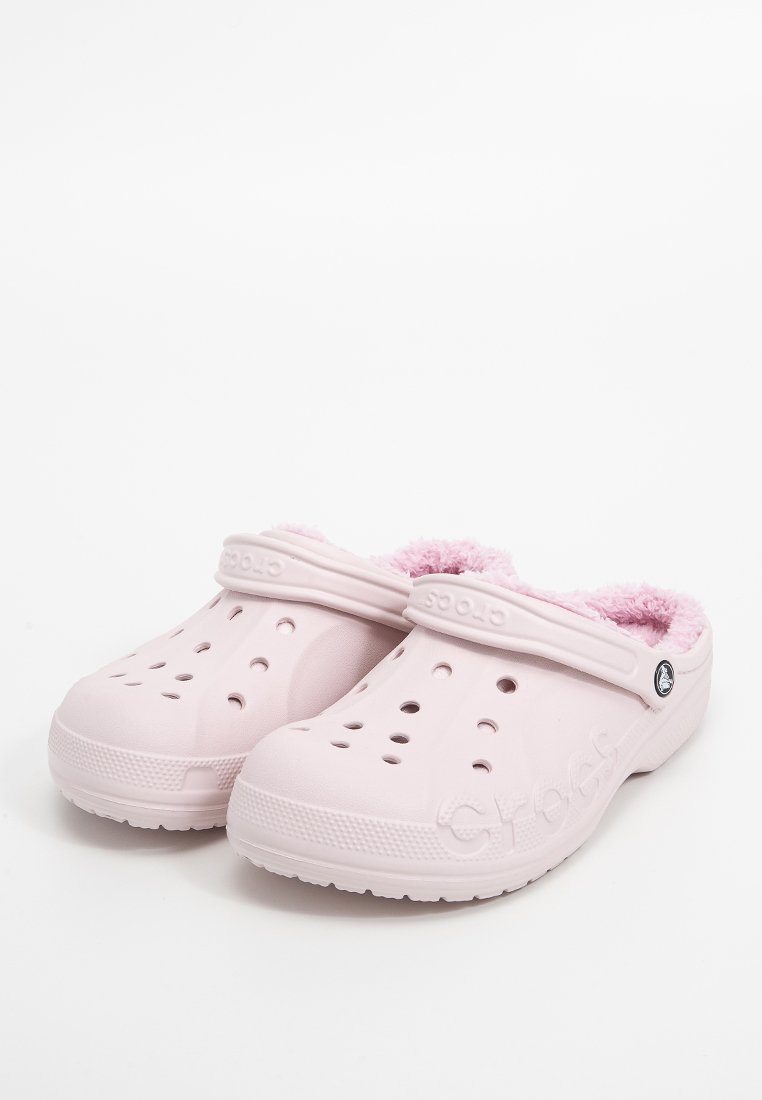 Zuecos de espuma rosa con forro de peluche, que cuentan con orificios de ventilación y una correa. Exterior texturizado y logotipo en el costado. Diseño ligero y casual.