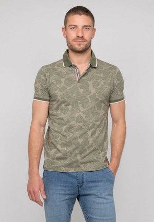 Homme aux cheveux courts et barbe portant un polo vert à motifs floraux et un jean bleu, debout avec une main dans la poche sur un fond uni.