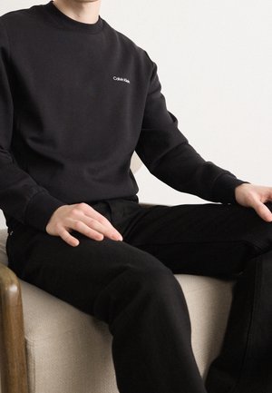 Person, der sidder på en beige stol iført en sort Calvin Klein-sweatshirt og sorte bukser med hænderne hvilende på benene.