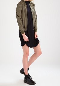 Oliivivihreä pommilentäjän takki mustan mekon päällä; istuva muotoilu, sileä materiaali, vetoketjukiinnitys, yhdistetty mustien kiiltävien nilkkabootsien kanssa.